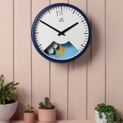 A round wall clock with a blue frame hangs on a pink paneled wall above a shelf with potted plants. The clock face features a mountain and sun illustration in the lower half.