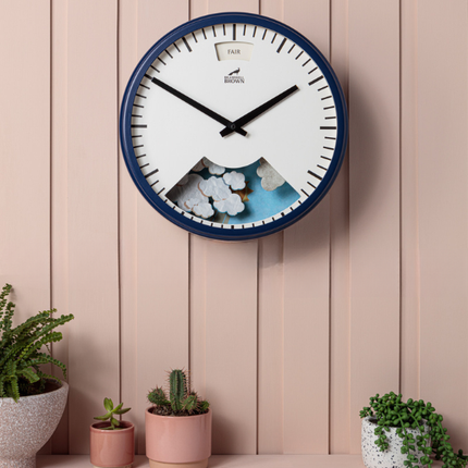 A round wall clock with a navy blue frame features a weather dial and a scene of clouds and mountains at the bottom. It hangs on a pink paneled wall above small potted plants, including succulents and cacti.