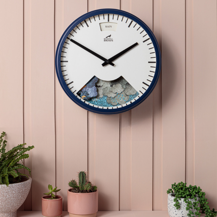A round wall clock with a navy blue rim and white face featuring a mountain design at the bottom, hanging on a light pink paneled wall above potted plants and succulents.