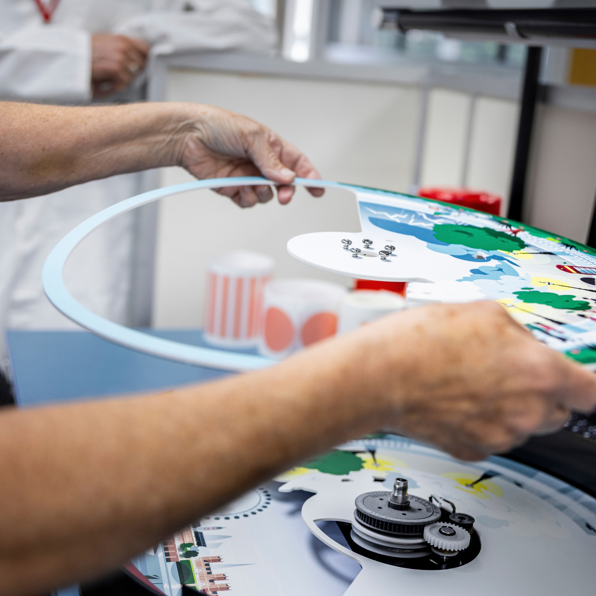 During a Clock Service and Refresh at the Workshop, hands carefully place a circular clock face illustration onto a mechanical movement.