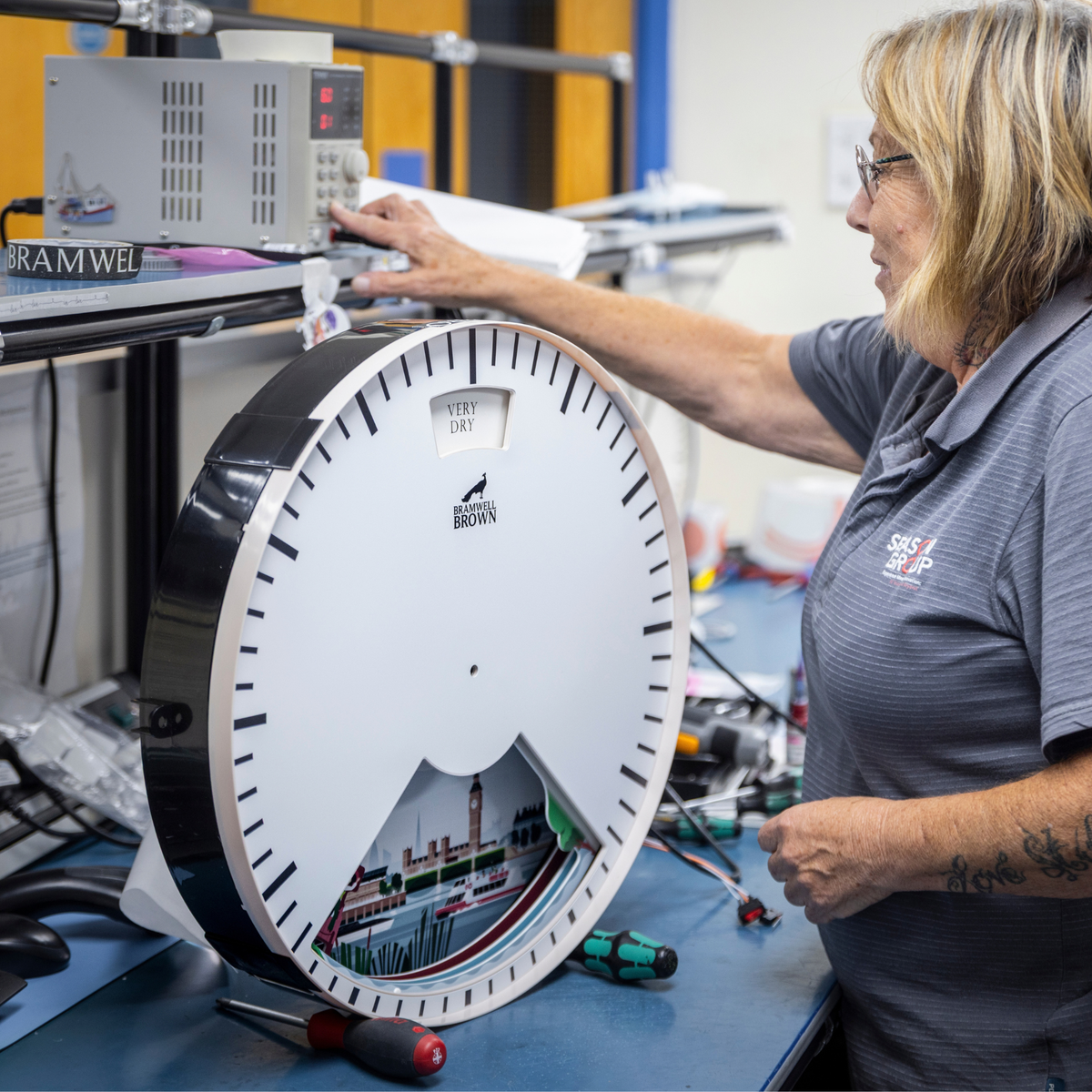 A person performs a Clock Service and Refresh at the Workshop on a large circular weather clock featuring a London cityscape illustration, preparing it for packaging on a workbench.