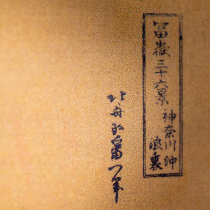 Close-up of Japanese calligraphy in black ink on a textured, tan-colored paper background. The text is arranged vertically in rectangular boxes and freeform, with no additional images or decorations.