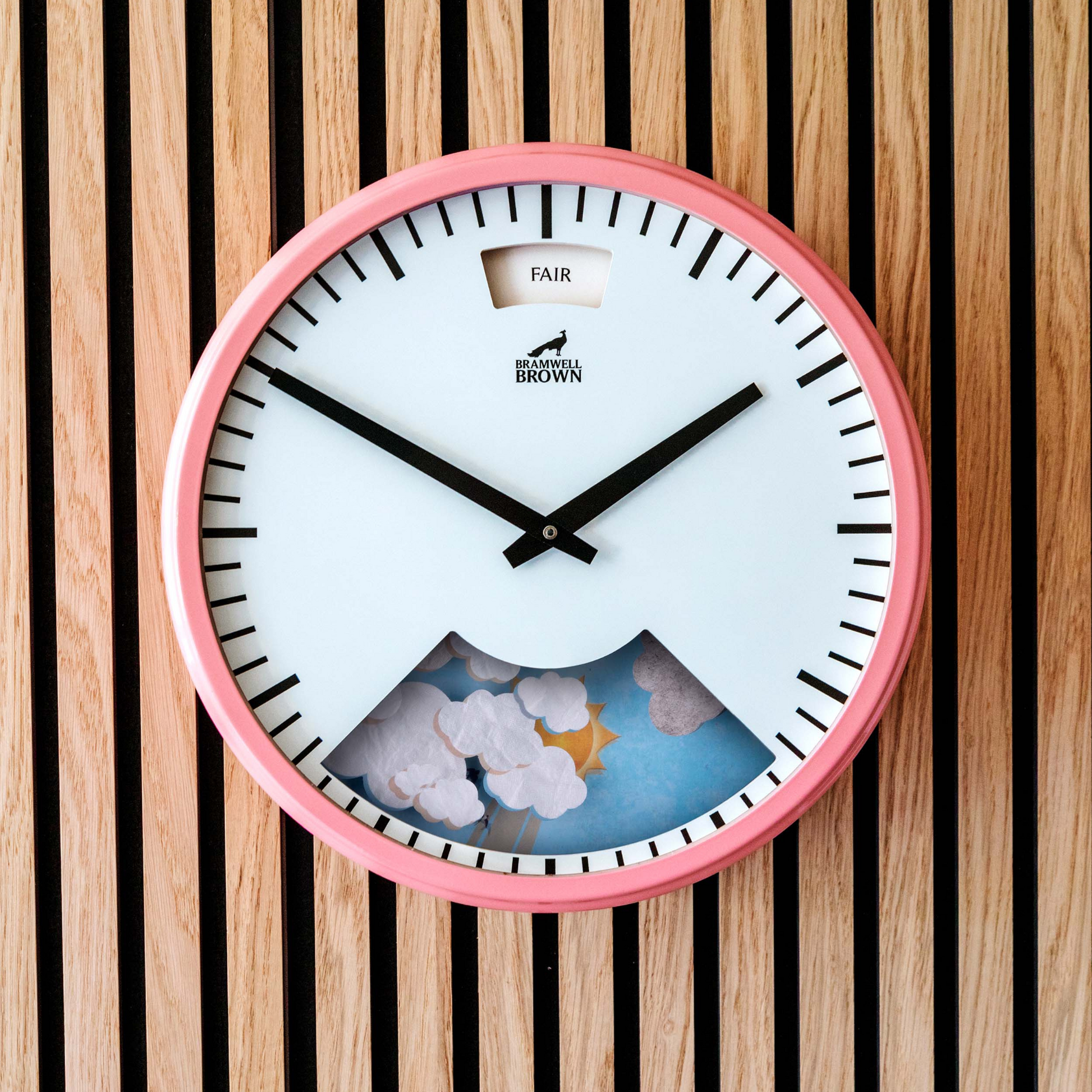 The Pink Weather Clock features a round, pink frame with black hands and hour marks. Its lower face displays clouds and a yellow star on light wood with vertical stripes, blending functionality with whimsical style.
