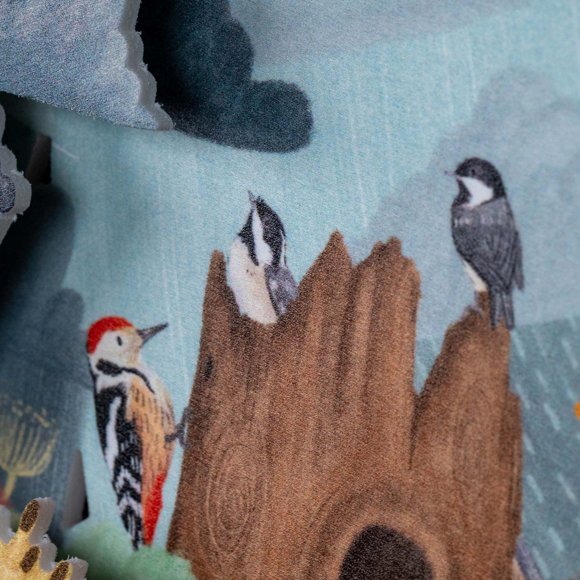 Illustration of three birds perched on a tree stump with blue sky and dark clouds in the background. One bird has a red head, while the others are grey and black. The scene is printed on a soft, textured fabric.