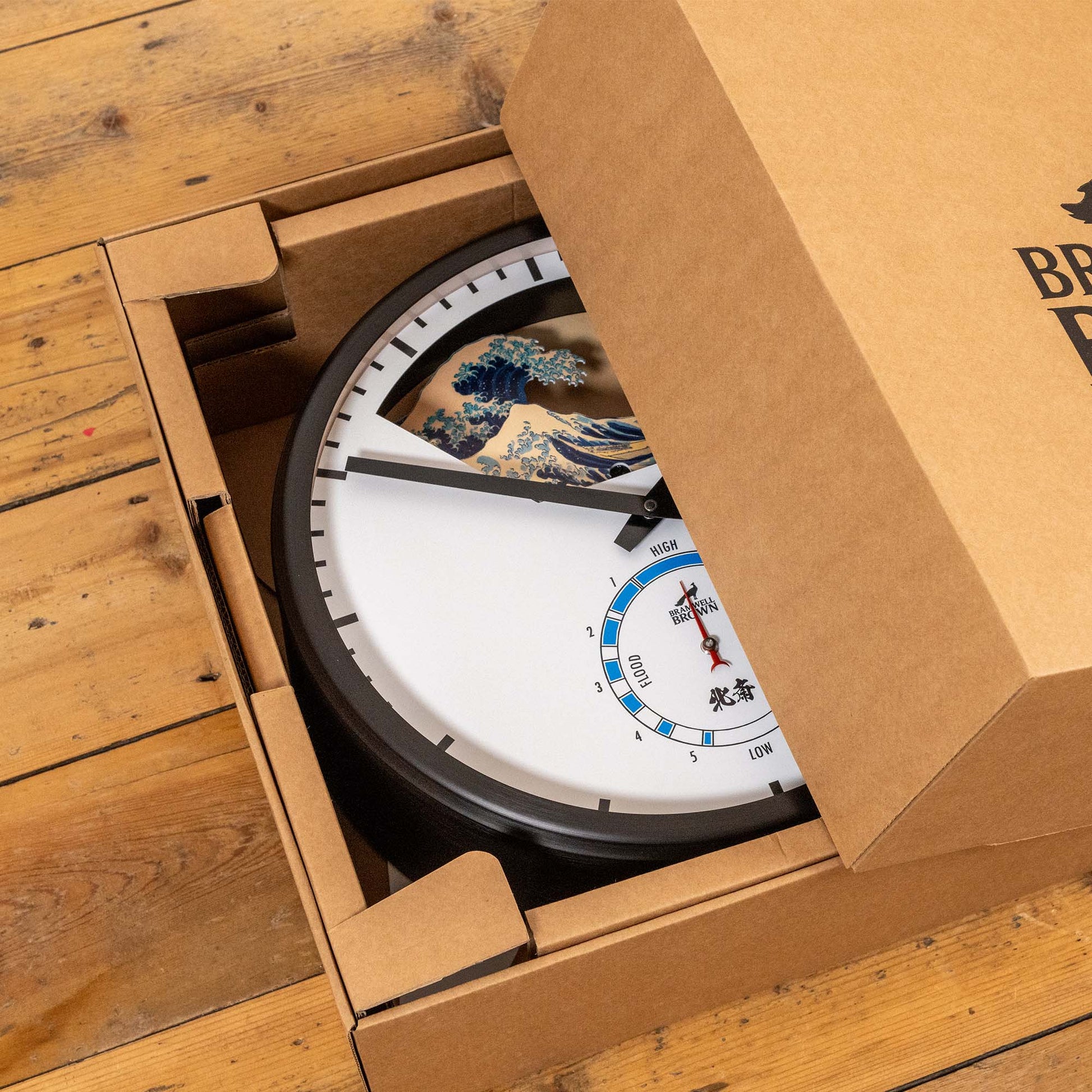 A partially unboxed Hokusai Tide Art Clock sits on a wooden floor. Its face showcases art inspired by The Great Wave Off Kanagawa and features a tide dial.