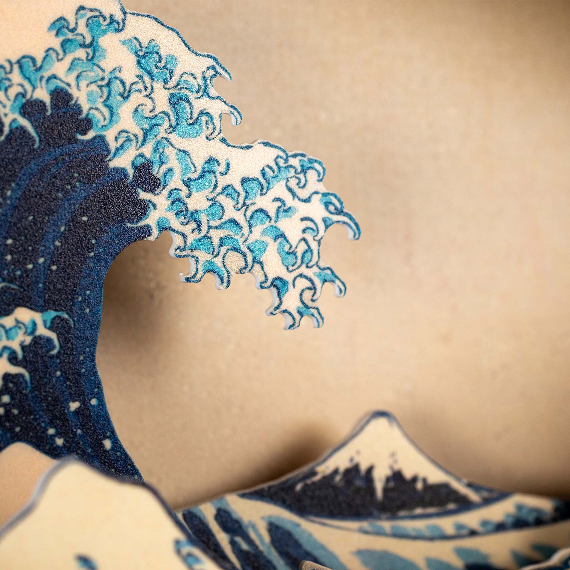Close-up of the Hokusai Tide Art Clock featuring a stylized blue and white wave inspired by the Great Wave Off Kanagawa, set against a beige background with part of a mountain at the bottom.