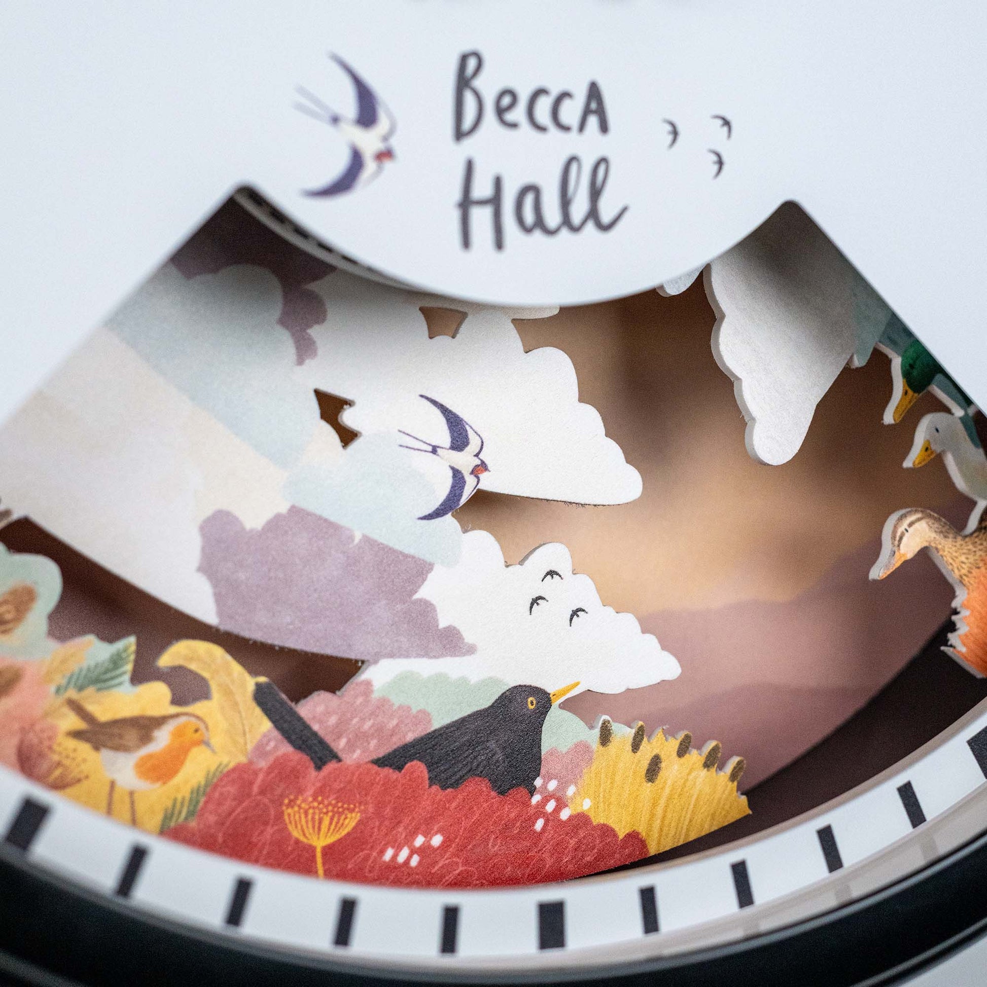 A close-up of a decorative paper diorama featuring colorful birds, clouds, and flowers, with Becca Hall handwritten above and small bird illustrations near the text.