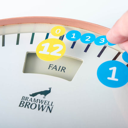 A hand places blue and yellow numbered stickers on a clock face labeled BRAMWELL BROWN. The clocks display reads FAIR. The numbers on the stickers are 0, 1, 2, 3, and 12.