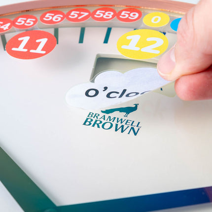 A hand peels a cloud-shaped sticker labeled Oclock from a Bramwell Brown clock face, with colorful numbered circles displayed along the top edge.