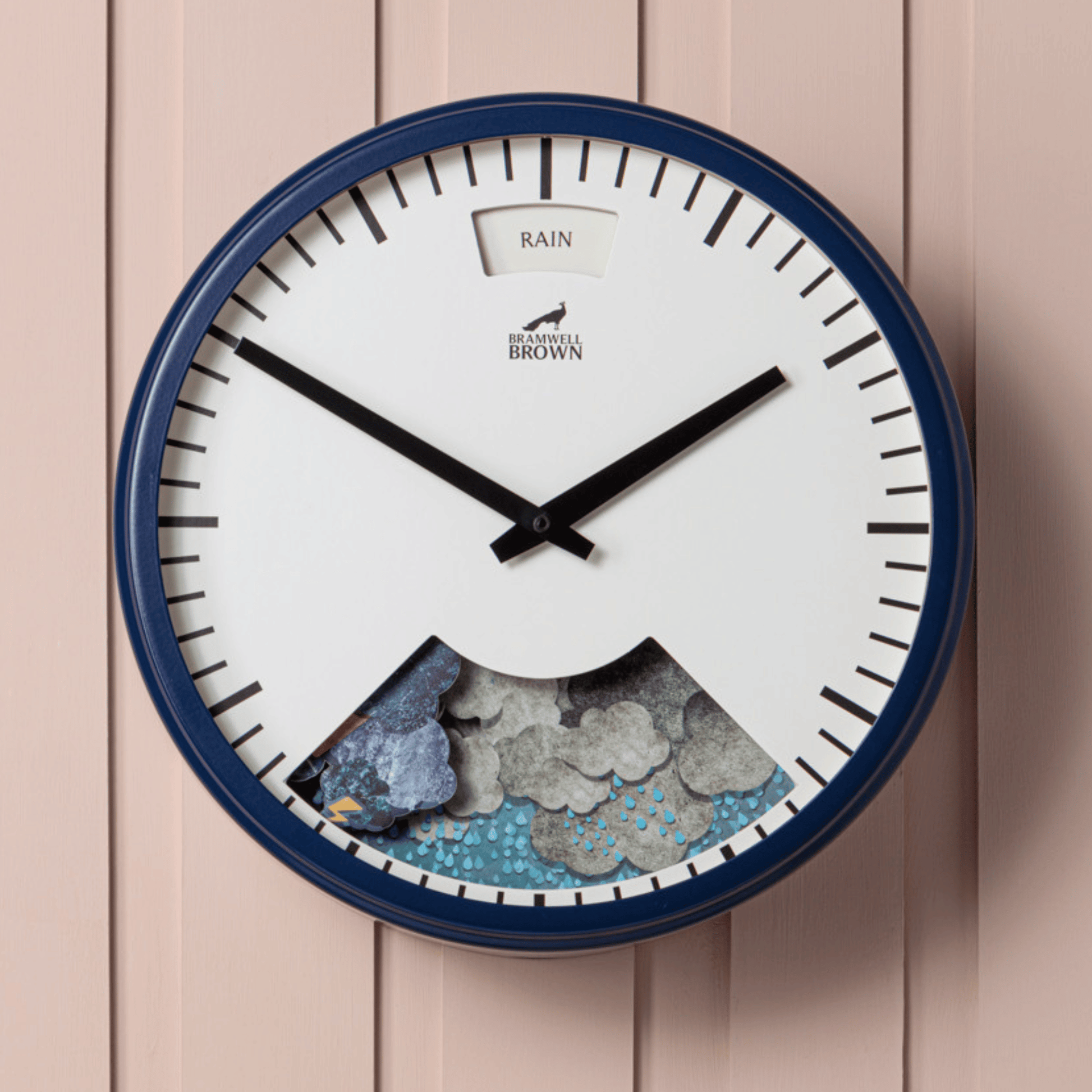 The Downpour Blue Weather Clock has a blue round frame, black hands, rain clouds and raindrops at the bottom, and a “RAIN” window at the top. This unique clock is mounted on pale pink paneling.
