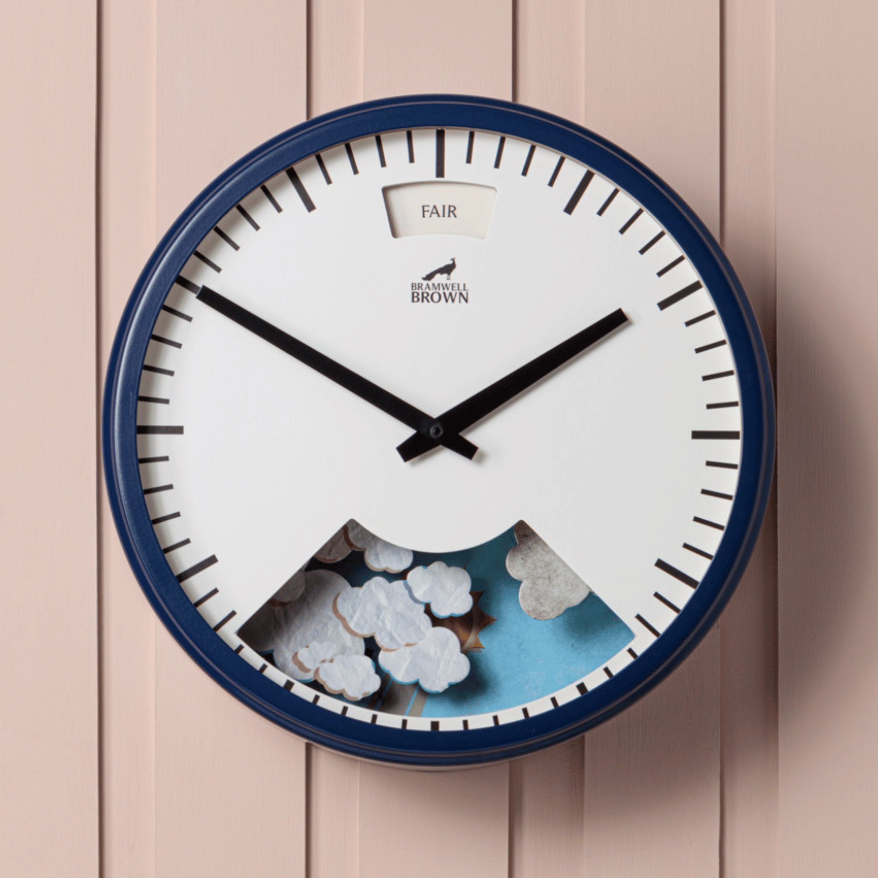 The Downpour Blue Weather Clock features black hands, a “FAIR” label, and a cutout showing clouds and sky. Hanging on light pink vertical paneling, this clock blends weather-inspired artistry with functional design.
