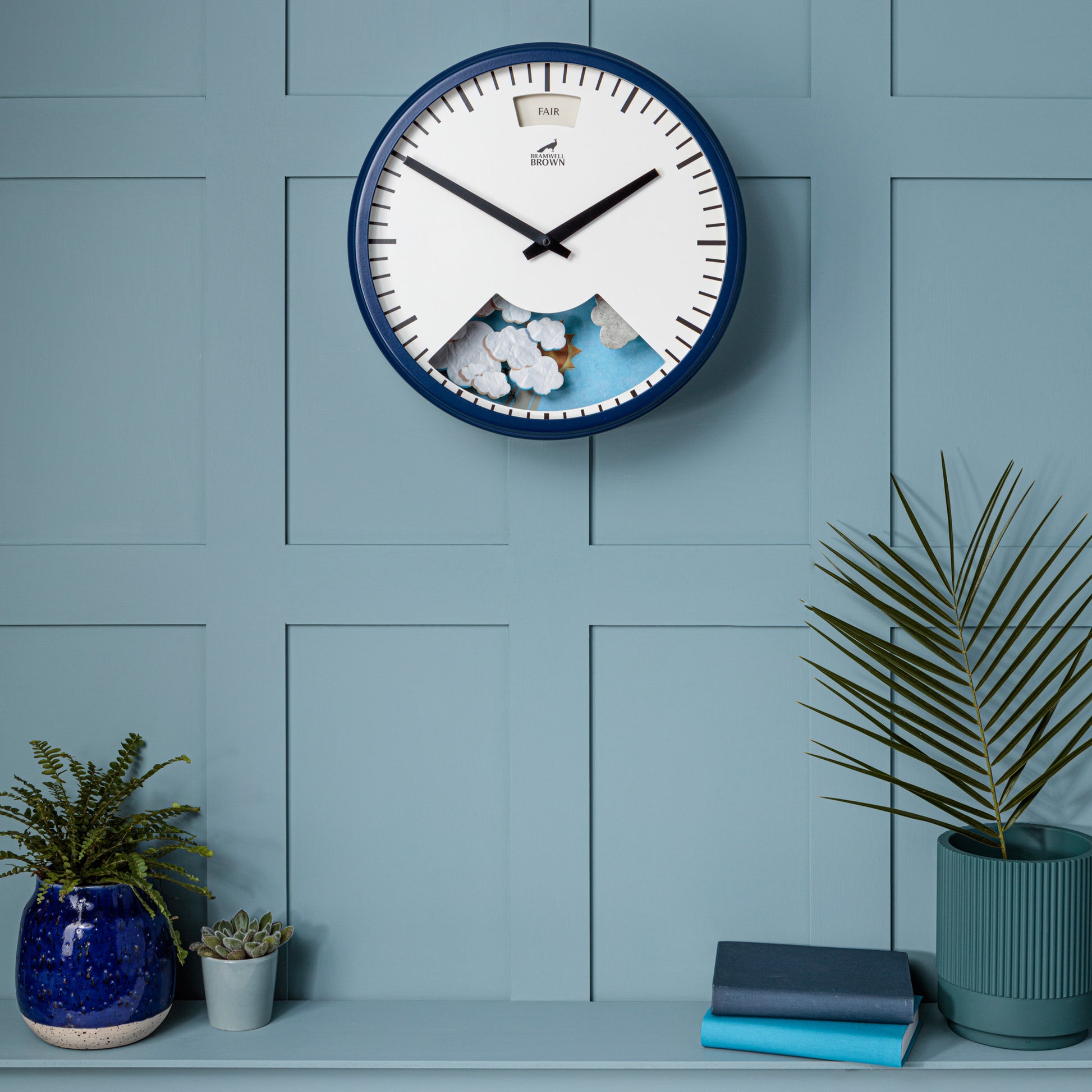 The Downpour Blue Weather Clock, featuring a sun and cloud design, hangs on a light blue paneled wall above a shelf with potted plants and books, highlighting its unique weather-inspired style and bespoke assembly.
