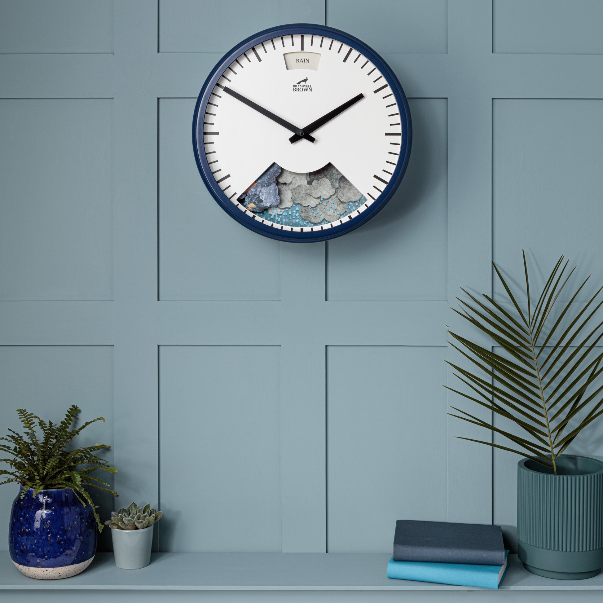 The Downpour Blue Weather Clock with black hands and no numbers hangs on a blue paneled wall above a shelf displaying a blue vase with a plant, a small potted plant, and two stacked books.