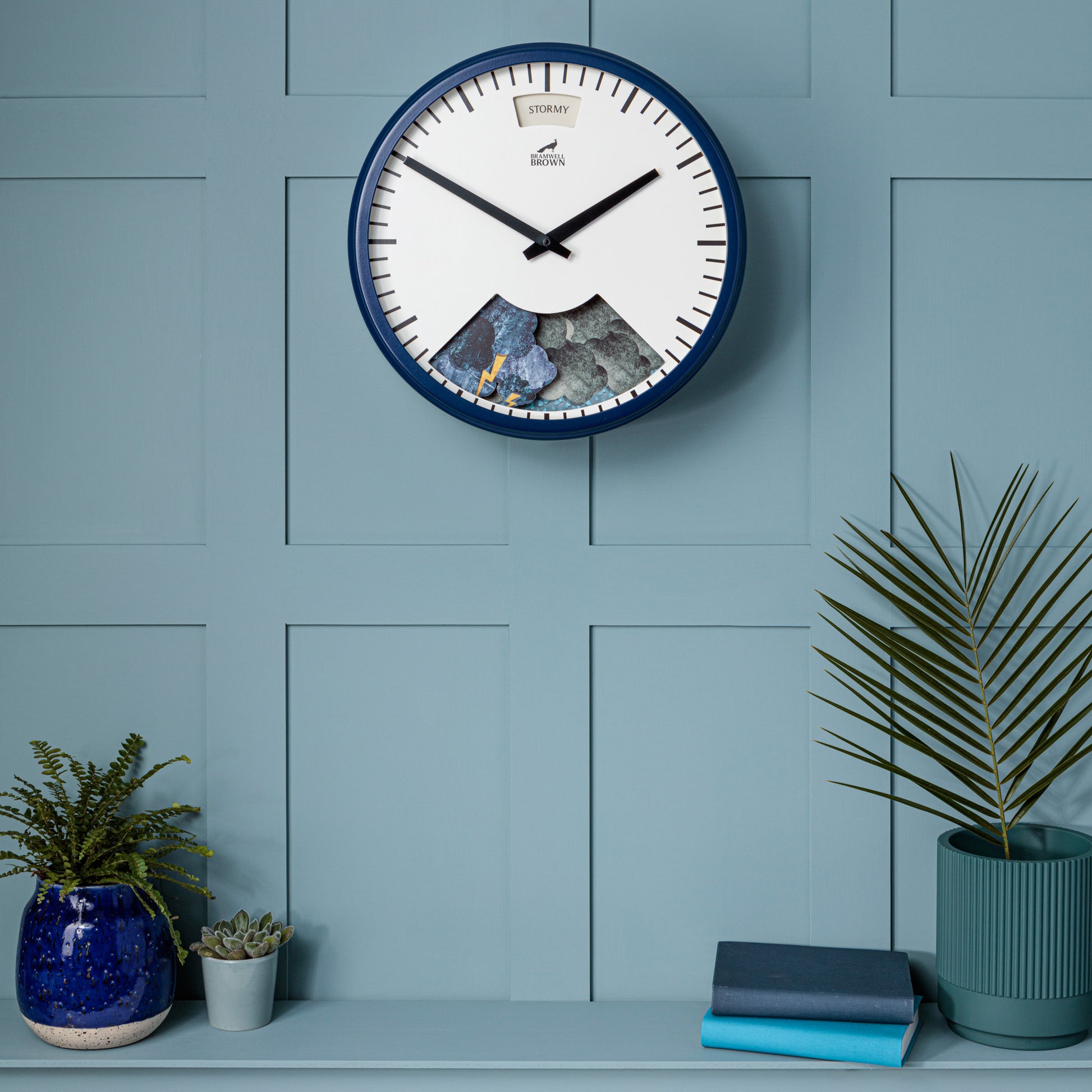 A Downpour Blue Weather Clock with a blue frame hangs on a light blue paneled wall above a shelf holding potted plants, a blue vase, and two closed books. The clock shows the time and features a stormy weather scene on its lower half.