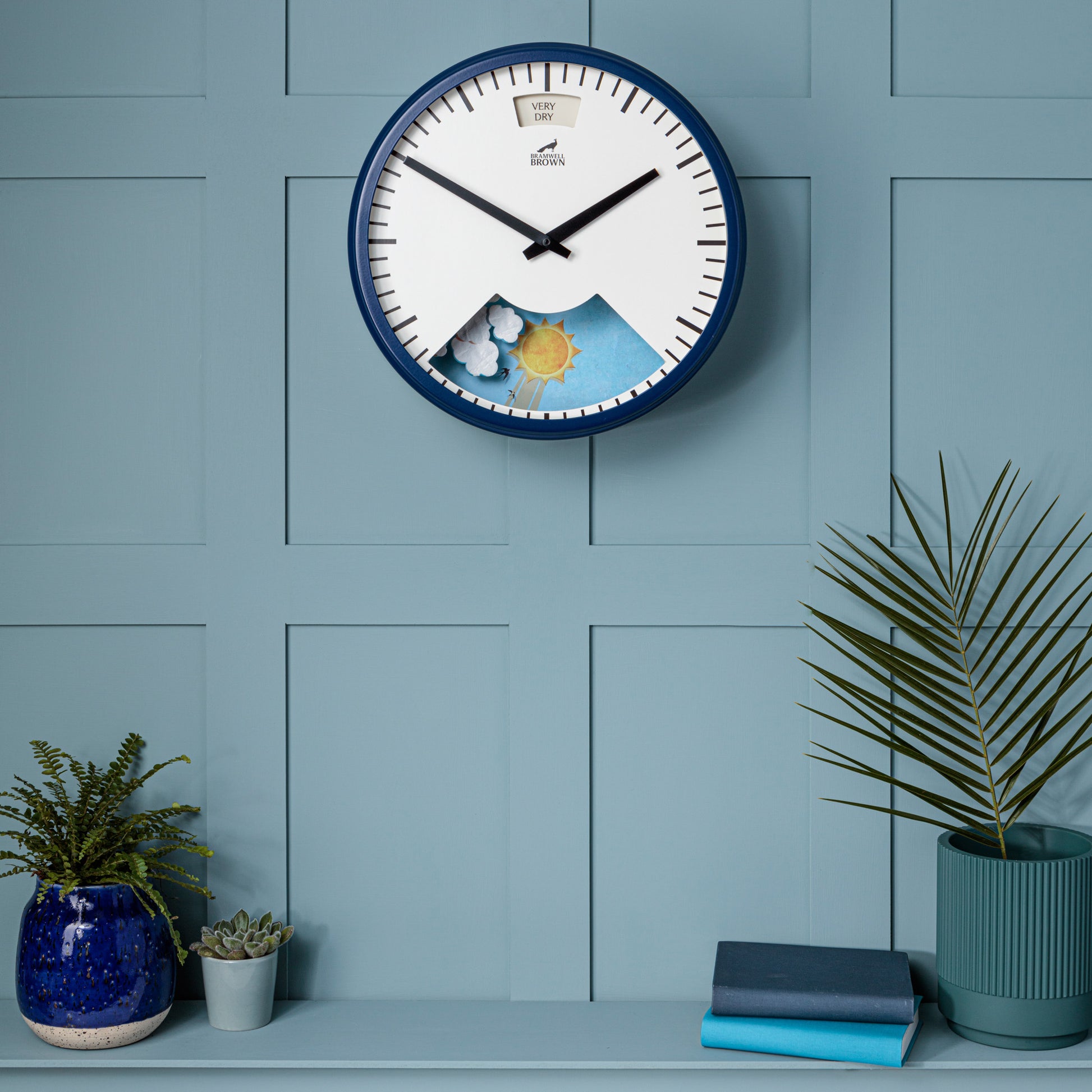 A Downpour Blue Weather Clock featuring a sun and clouds illustration hangs on a light blue paneled wall above a shelf with potted plants, a blue vase, and two closed books.