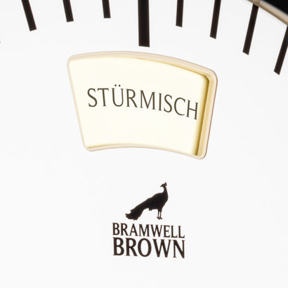 Close-up of the German-language Weather Clock’s white dial, displaying the word STÜRMISCH in a gold-framed window above the Bramwell Brown logo, featuring a peacock silhouette. Inspired by traditional German weather forecasting, this unique clock combines artistry and precision—making it an exceptional piece of wall art. Black tick marks are visible at the top of the image.