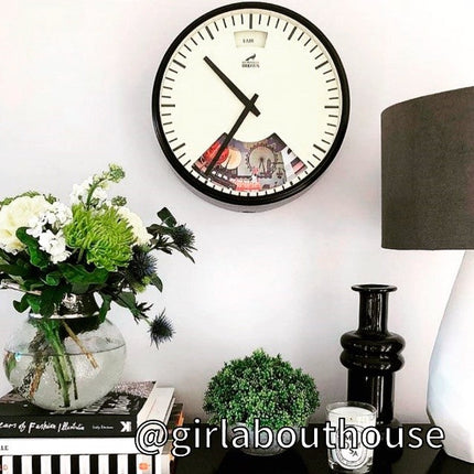 A round wall clock with black rim hangs above a table with a vase of flowers, a small green plant, a black vase, a candle, stacked books, and a gray lampshade. @girlabouthouse is written at the bottom.