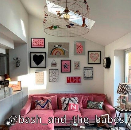 A bright living room with a pink sofa, colorful cushions, and framed art on the wall. The artwork includes phrases, hearts, a rainbow, and geometric patterns. A large wire lampshade hangs from the ceiling.