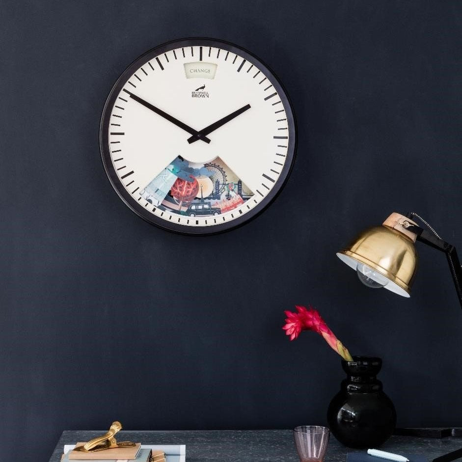 A round wall clock with a white face and black frame; part of the lower half is cut out, revealing colorful banknotes and coins inside. Below, a desk holds a gold lamp, red flower in a vase, and various stationery items.