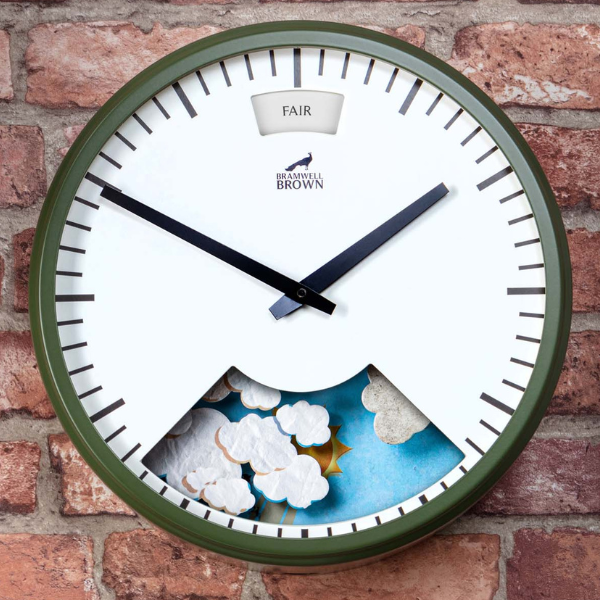 An Olive Green Weather Clock, with black hands and an olive green frame, displays weather information—showing “FAIR” at the top and featuring sun and clouds in a cut-out at the bottom. This distinctive weather clock is mounted on a brick wall.
