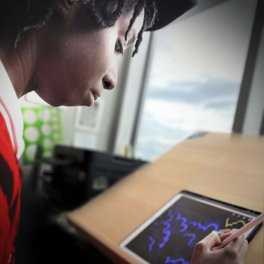 A person wearing a red shirt and cap is drawing colorful designs on a tablet with a stylus while sitting at a wooden desk near a window with a cloudy sky visible outside.