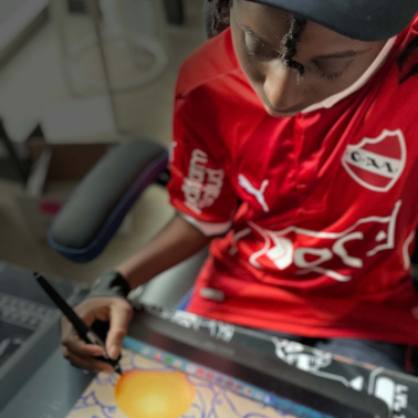 A person wearing a red soccer jersey and cap draws a digital illustration of an orange fruit on a tablet, using a stylus, while seated at a desk.