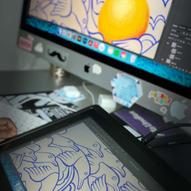 A digital artist works on a tablet connected to a desktop computer, illustrating blue bird outlines and a large yellow sun. The workspace is decorated with stickers and colorful items.