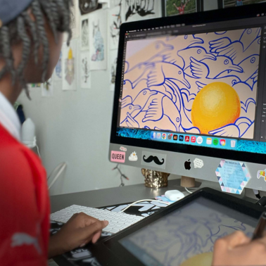 A person uses a digital drawing tablet at a desk, illustrating birds and a large yellow circle on a computer screen. The workspace is decorated with art and stickers, including one that says QUEEN.