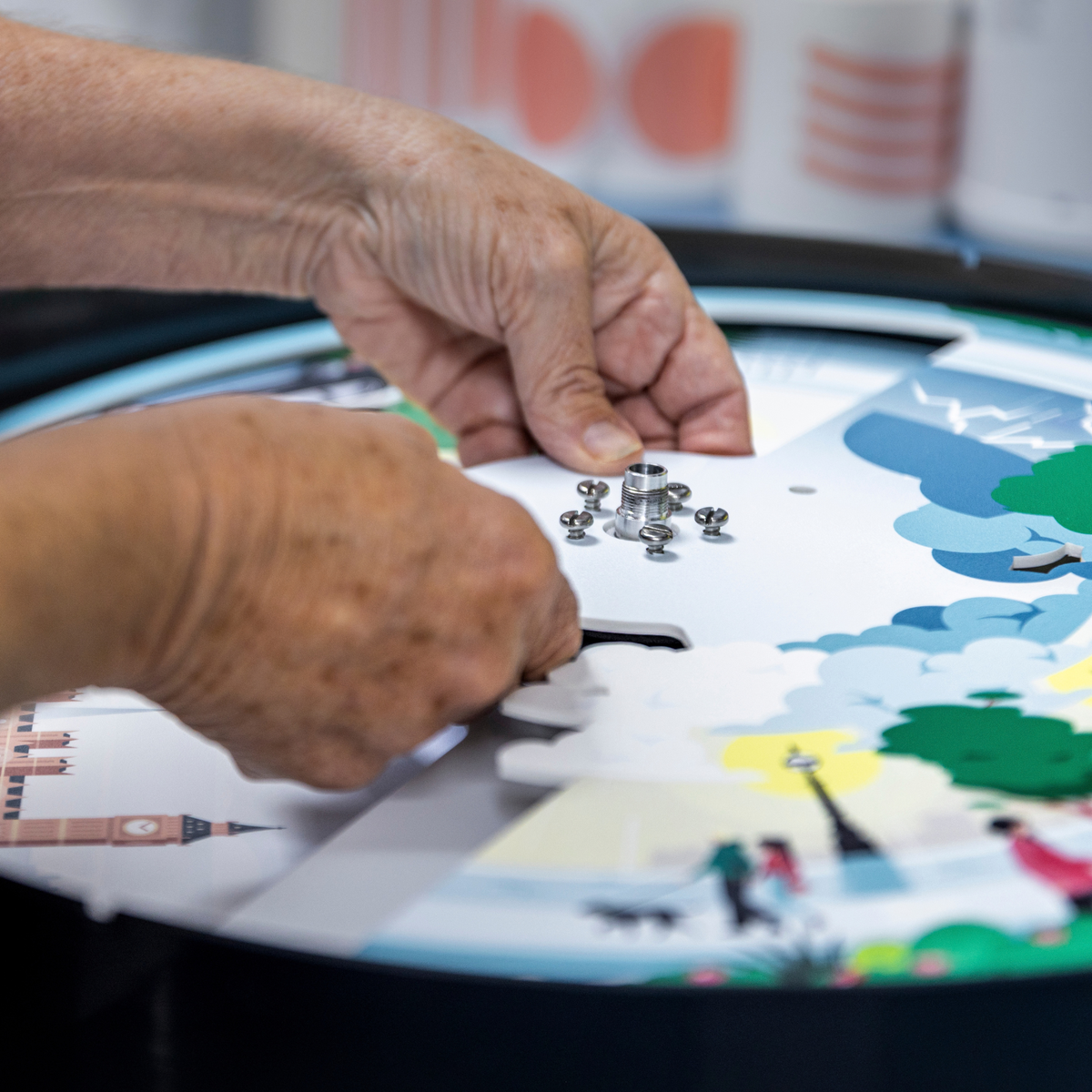 Hands place a clock mechanism onto an illustrated clock face during Clock Service and Refresh at the Workshop.