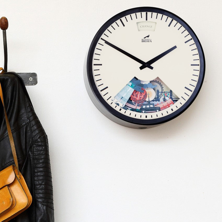 A wall clock with a clear section at the bottom displaying assorted coins and bills. The clock hangs above a coat hook holding a black leather jacket and a tan handbag against a white wall.