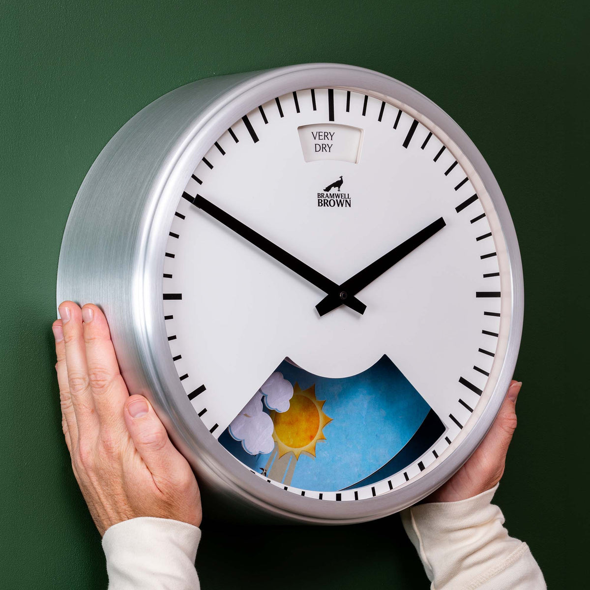 A person holds a Chrome framed Weather Art Clock—featuring black hands, tick marks, weather symbols at the bottom, and the text “VERY DRY” near the top—against a green wall.