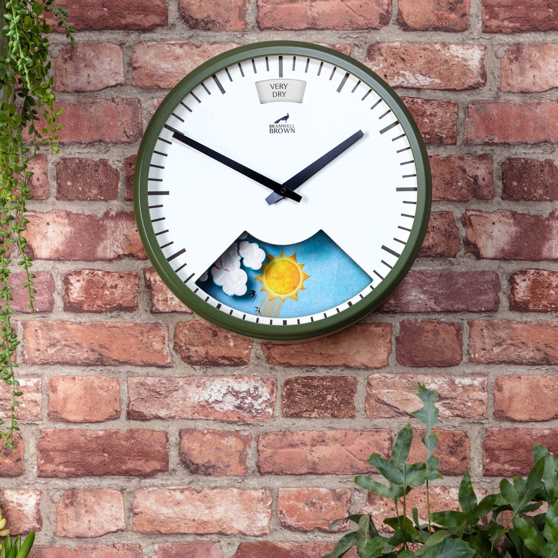 The Olive Green Weather Clock features black hands and is mounted on a brick wall, displaying a sunny weather scene at the bottom. Green plants frame parts of the image, complementing this unique and stylish décor piece.
