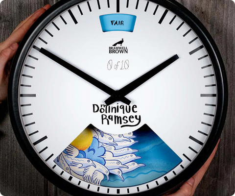 A round wall clock with bold black hands, a white face, and a “Bramwell Brown” logo. The lower section displays animated blue-and-white birds flying. The word FAIR appears at the top and Dominique Ramsey is written in the center.
