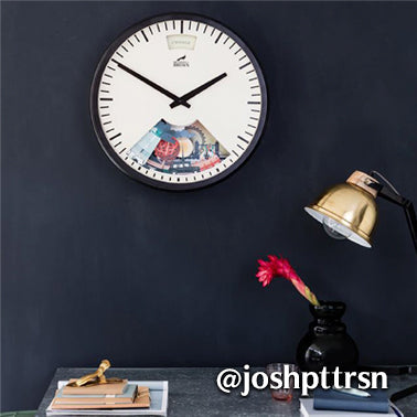 A wall clock with a black frame and white face displays two black hands, with a small section showing a collage of images. Below, a desk holds books, a gold lamp, a vase with a red flower, and various small items.