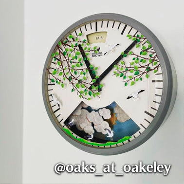 Round wall clock with black hands, decorated with green-leafed branches, white birds, and clouds. The upper part displays a FAIR weather indicator. Instagram handle @oaks_at_oakeley is overlaid at the bottom.