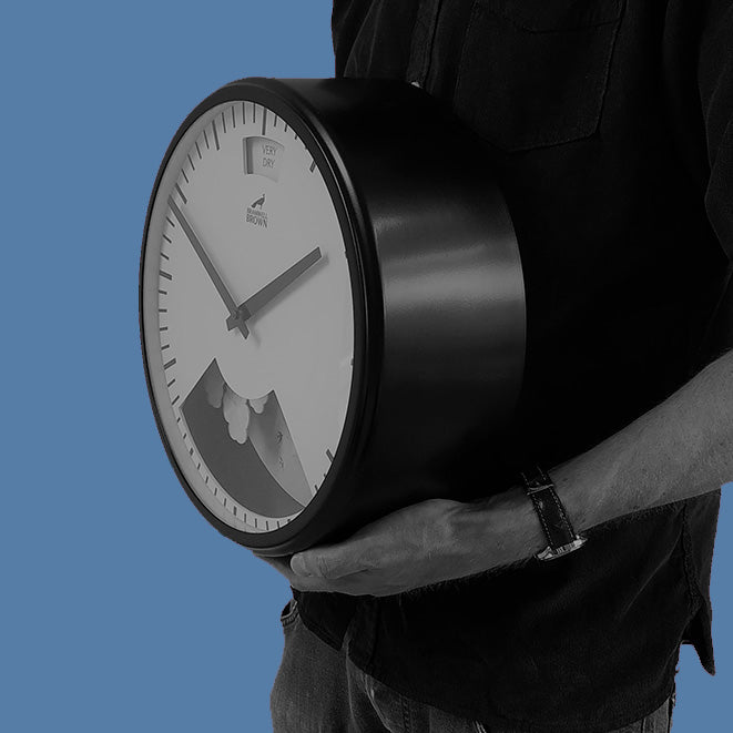 A person in a dark shirt holds a large round clock with a black frame and a white face, showing only one hand. The background is a solid blue color.