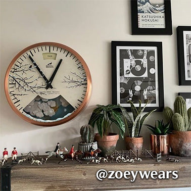 A wall with a large clock featuring a mountain and cherry blossom design, framed art, potted cacti, and miniature horse and rider figurines arranged on a wooden shelf. @zoeywears is written on the image.
